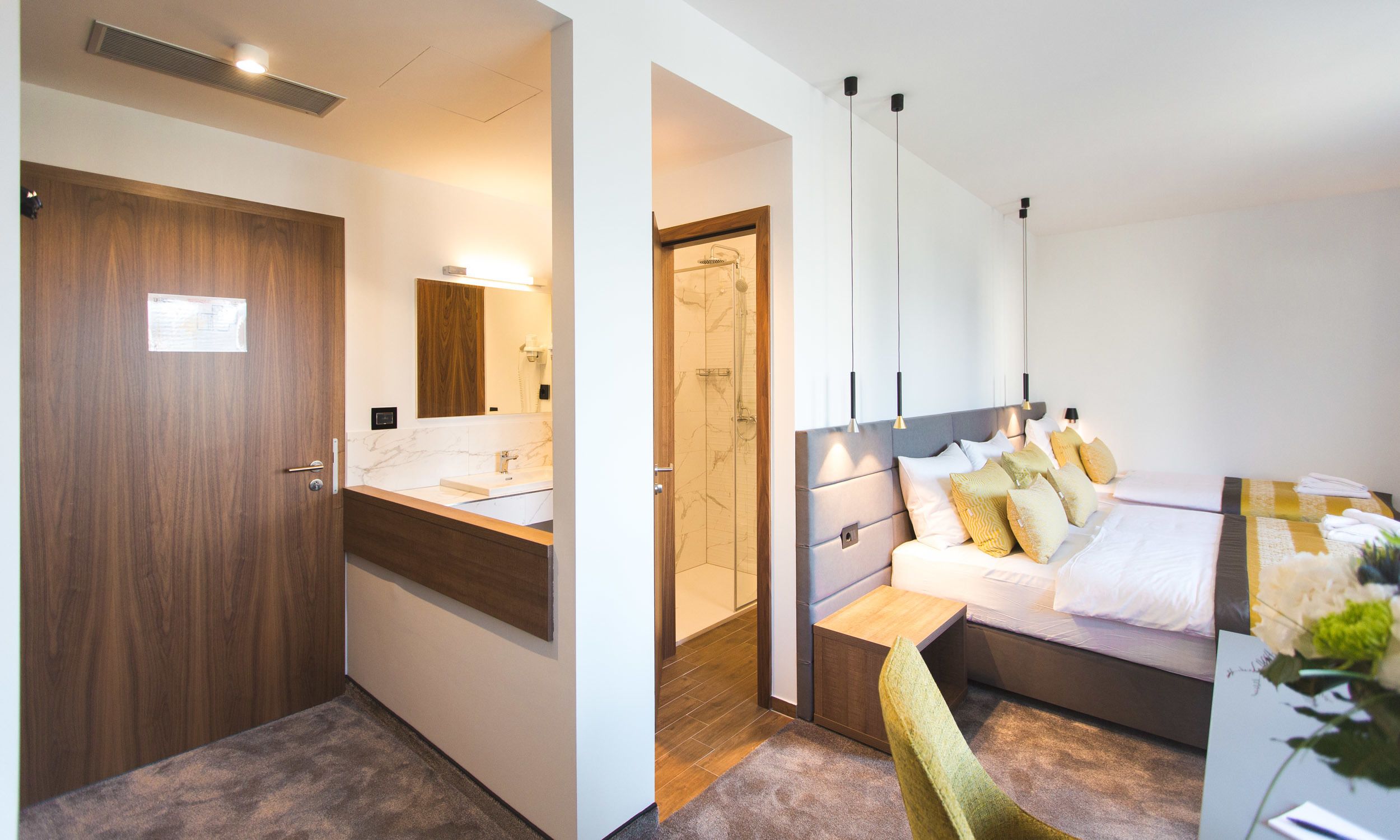 A hotel room with a wooden door, a bathroom with a mirror, a shower, a bed with multiple pillows, and a bedside table.