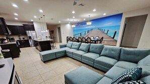 A modern living room with a large teal sectional sofa, a dining area with a wooden table and chairs, and a kitchen with dark cabinets and stainless steel appliances. - Shv1206ha - 8 Bedroom Villa In Windsor At Westside, Sleeps Up To 18, Just 7 Miles To Disney (Kissimmee)
