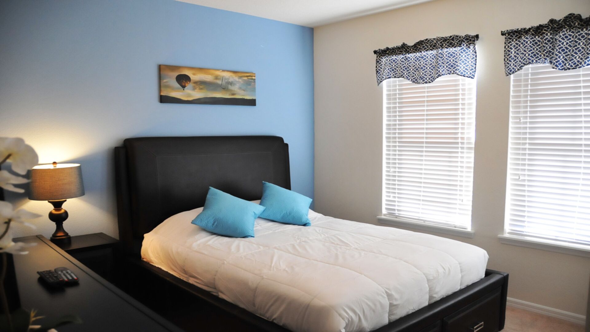A bedroom with a bed, a nightstand with a lamp, and two windows with patterned curtains.