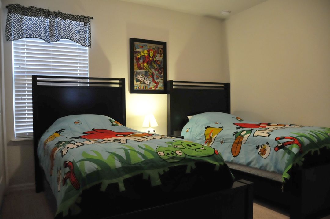Une chambre avec deux lits, chacun recouvert d’une couette aux motifs de dessins animés, une fenêtre avec des stores et un tableau encadré au mur.