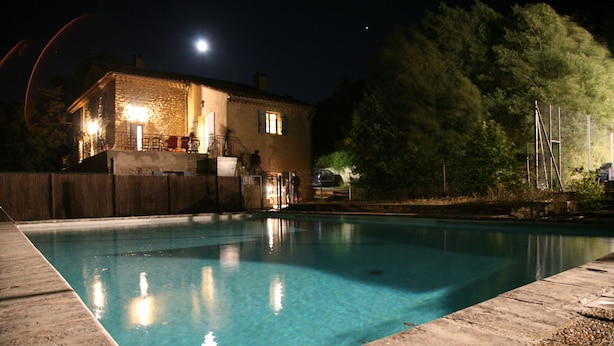 A swimming pool with a stone border, a stone building with a balcony, and a car parked nearby. - Maison de Veaux (Malaucène)