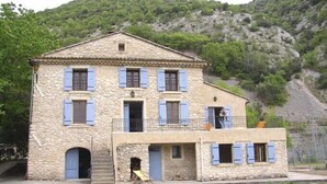A two-story stone building with blue shutters, a balcony, and a small attached structure with a door and window. - Maison de Veaux (Malaucène)