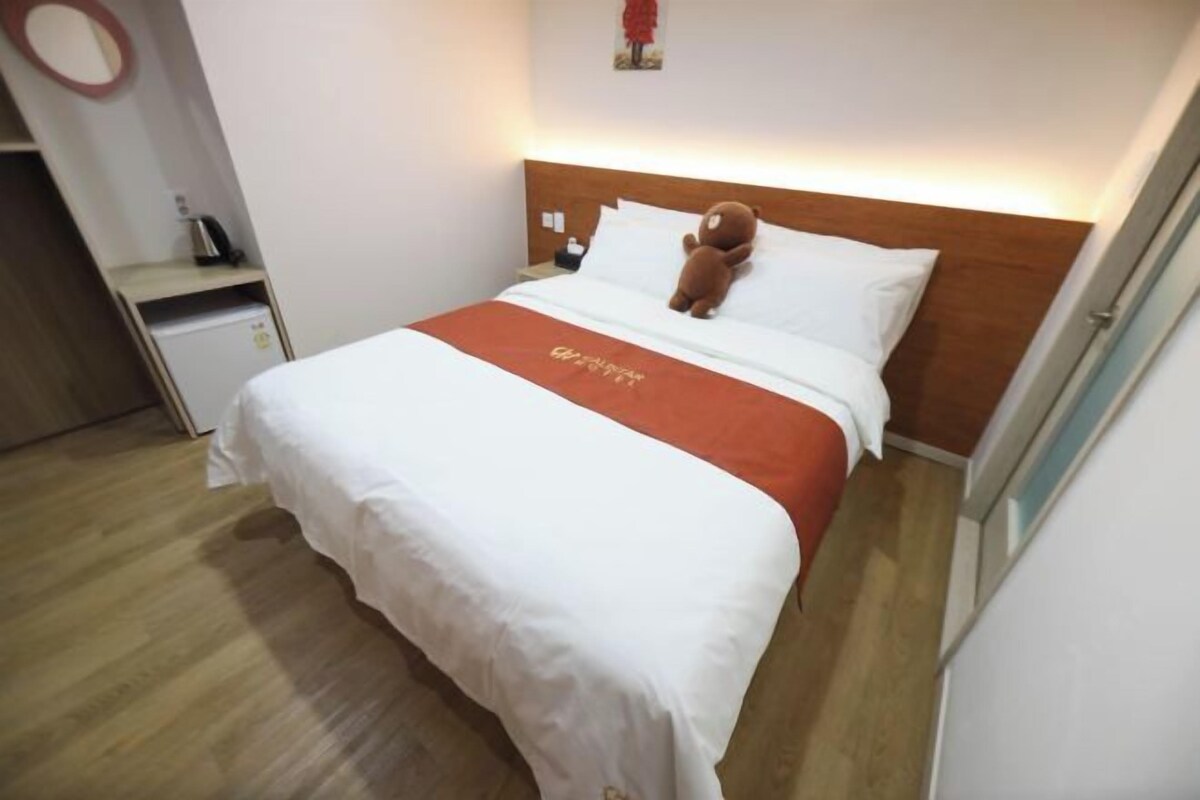 A hotel room with a bed, a teddy bear on the pillows, a small table with a kettle, and a wall-mounted mirror.