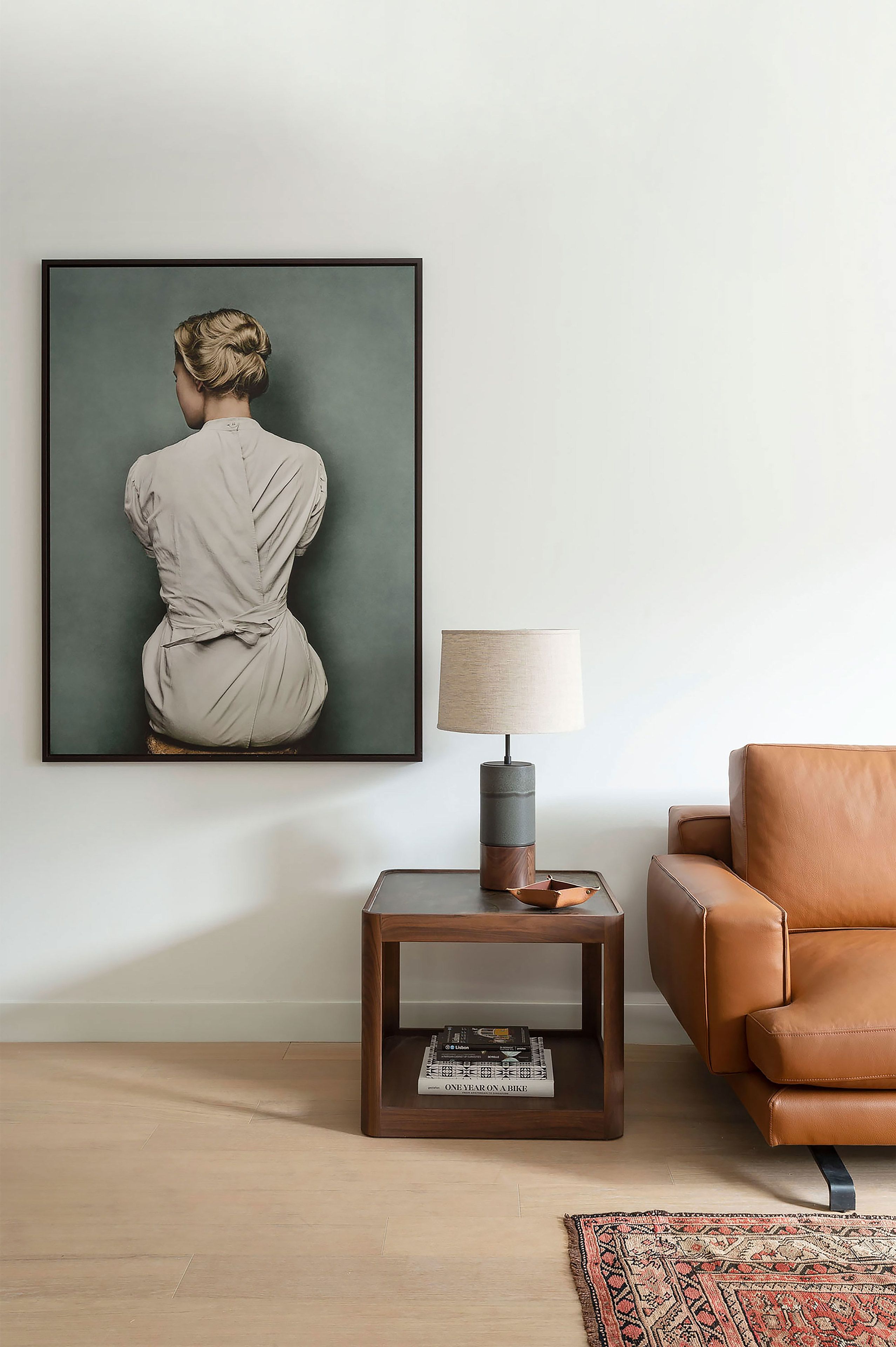Una obra de arte enmarco que muestra una figura sentada, una mesita de madera con una lámpara, un sillón de cuero y una alfombra estampada sobre un piso de madera.