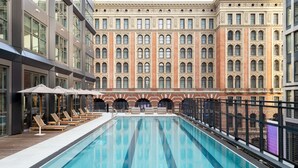 Ein Pool auf dem Dach mit Liegestühlen und Sonnenschirmen, umgeben von einem historischen Gebäude mit Rundbogenfenstern und einer Ziegelaußenwand.