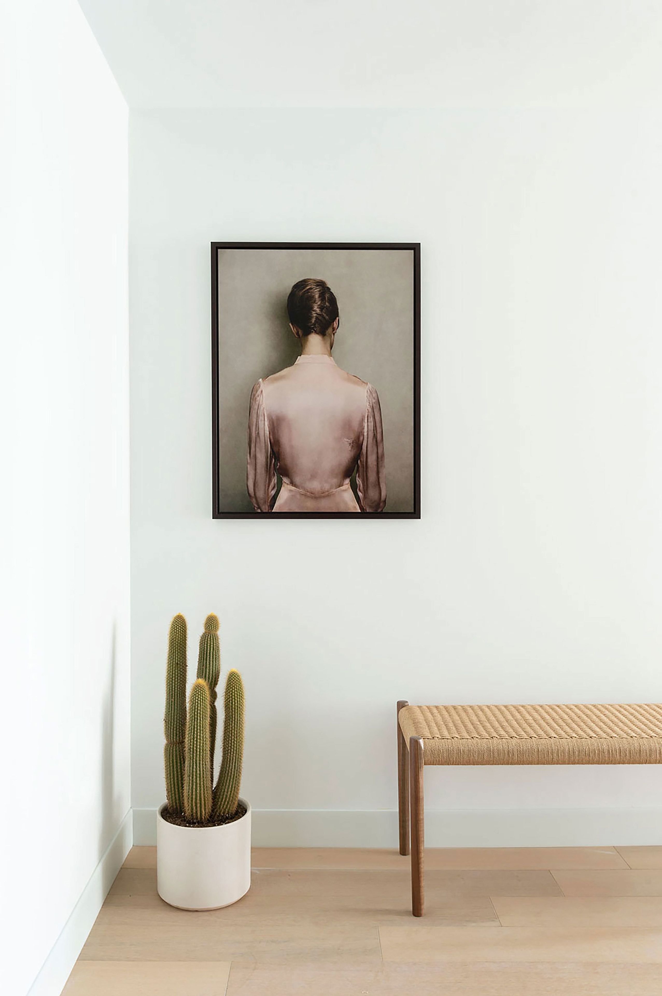 A framed artwork of a person from behind, a potted cactus, and a woven bench against a white wall.