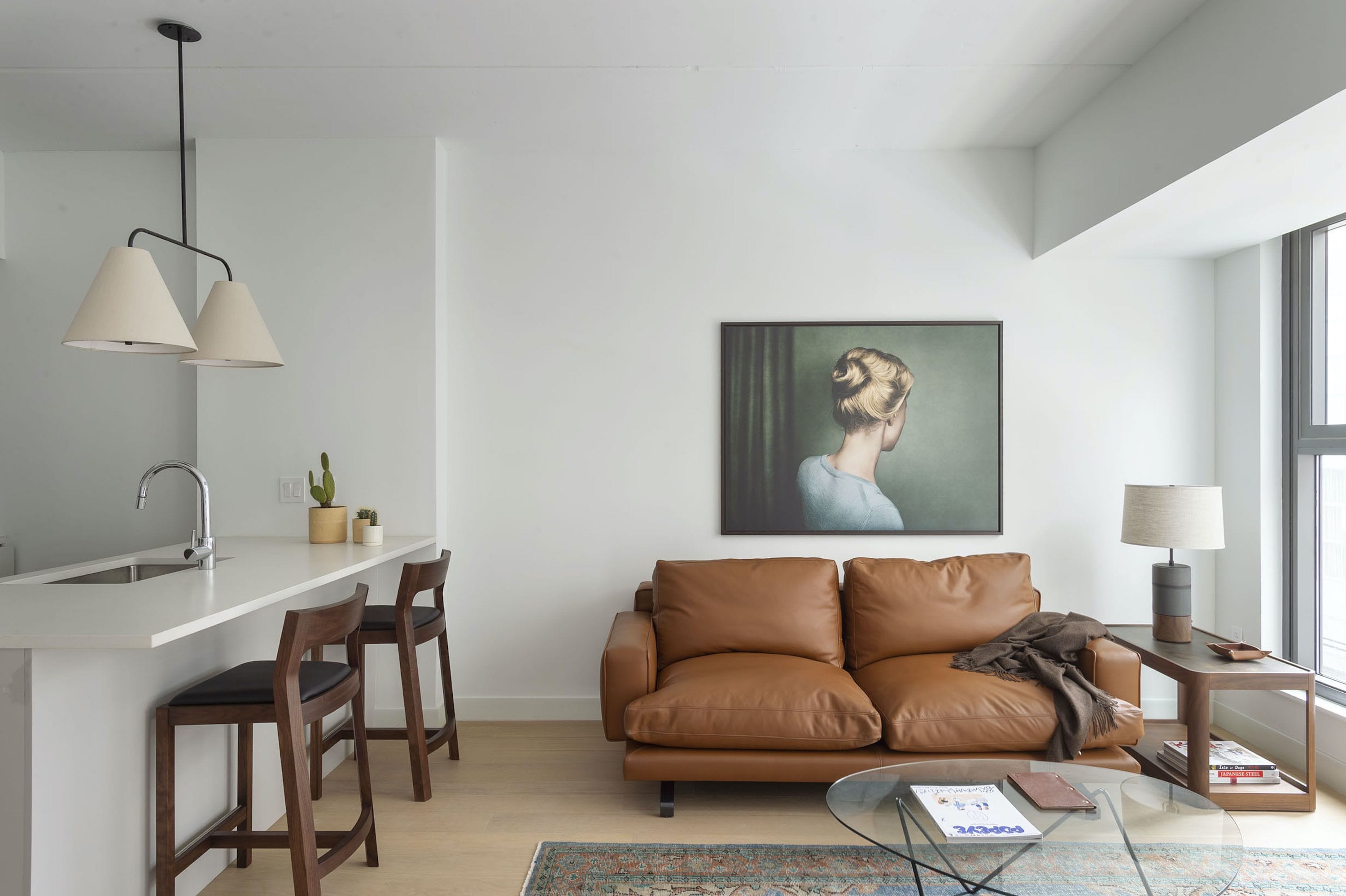 Un espace de vie moderne comprenant un canapé en cuir marron, une table basse en verre, un coin cuisine blanc avec un évier et une grande fenêtre laissant entrer la lumière naturelle.