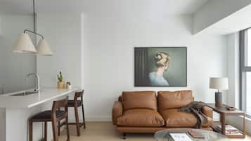 Un espace de vie moderne comprenant un canapé en cuir marron, une table basse en verre, un coin cuisine blanc avec un évier et une grande fenêtre laissant entrer la lumière naturelle.