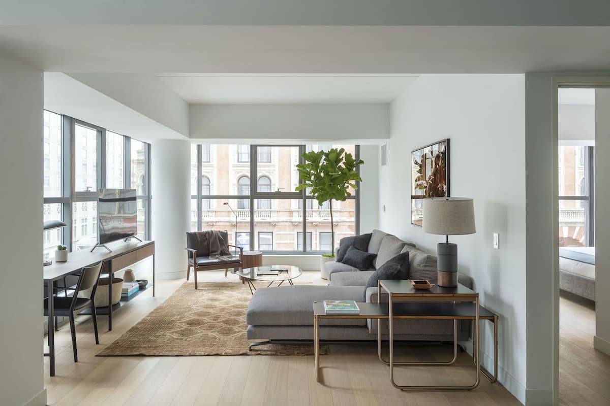 A modern living room with a sofa, coffee table, and a desk with a computer. Large windows provide a view of the city.