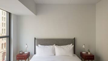 Quarto de hotel moderno com uma janela grande, uma cama com roupa branca, duas mesas de cabeceira com abajures e vista para um edifício.