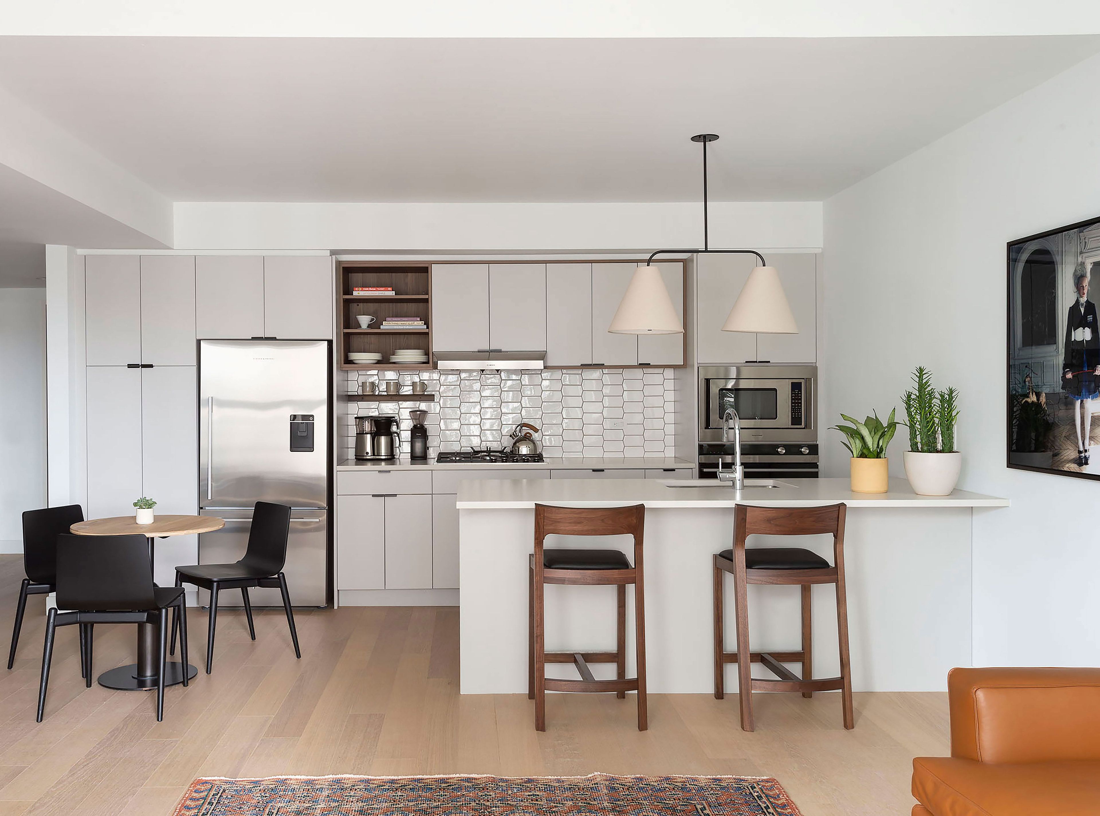Una cocina moderna con armarios blancos, un refrigerador de acero inoxidable, un microondas y una estufa a gas. Hay dos taburetes de bar de madera y una pequeña mesa redonda con sillas.