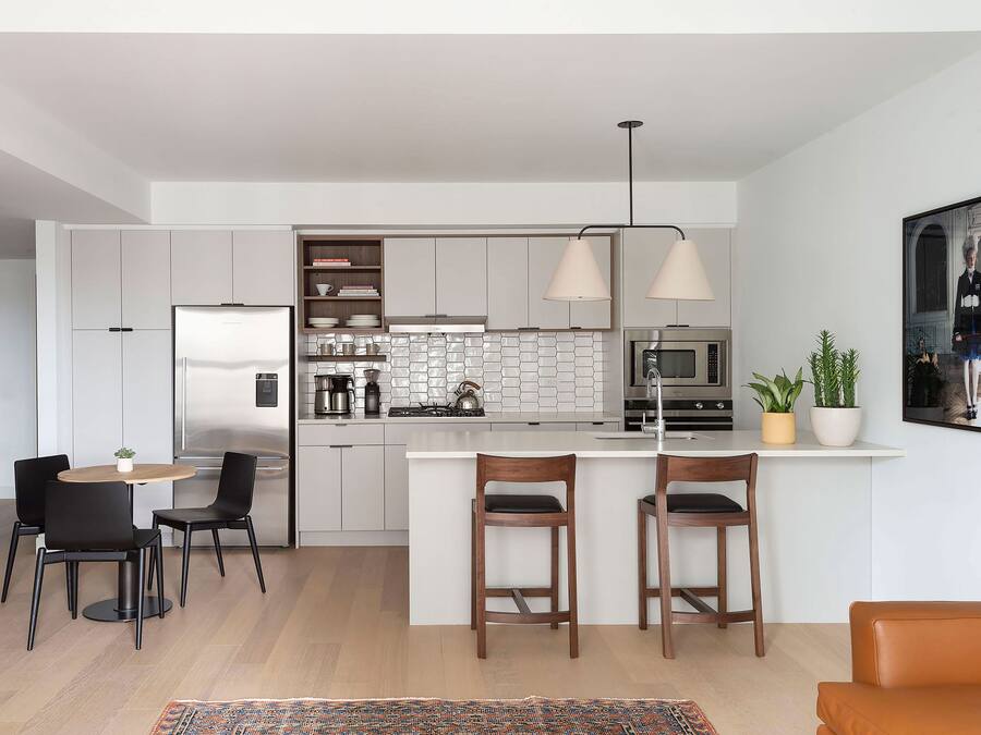 Een moderne keuken met witte kasten, een roestvrijstalen koelkast, een magnetron en een gasfornuis. Er zijn twee houten barstoelen en een kleine ronde tafel met stoelen.
