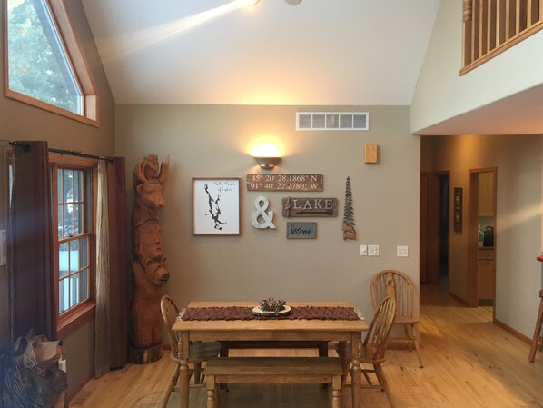 A dining area with a wooden table and chairs, a large window with curtains, and wall decorations including a map and a 'Lake' sign. - Sunny's Landing 3000 SF home on Prairie Lake (Chetek)