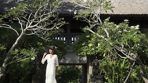 A woman in a white dress and hat walking on a pathway in a garden with a thatched-roof structure in the background. - REVĪVŌ Wellness Resort Nusa Dua Bali (Nusa Dua)