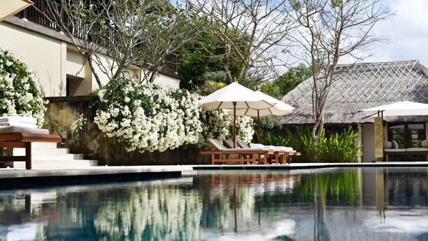 A pool area with lounge chairs and umbrellas, surrounded by lush greenery and white flowering plants. - REVĪVŌ Wellness Resort Nusa Dua Bali (Nusa Dua)