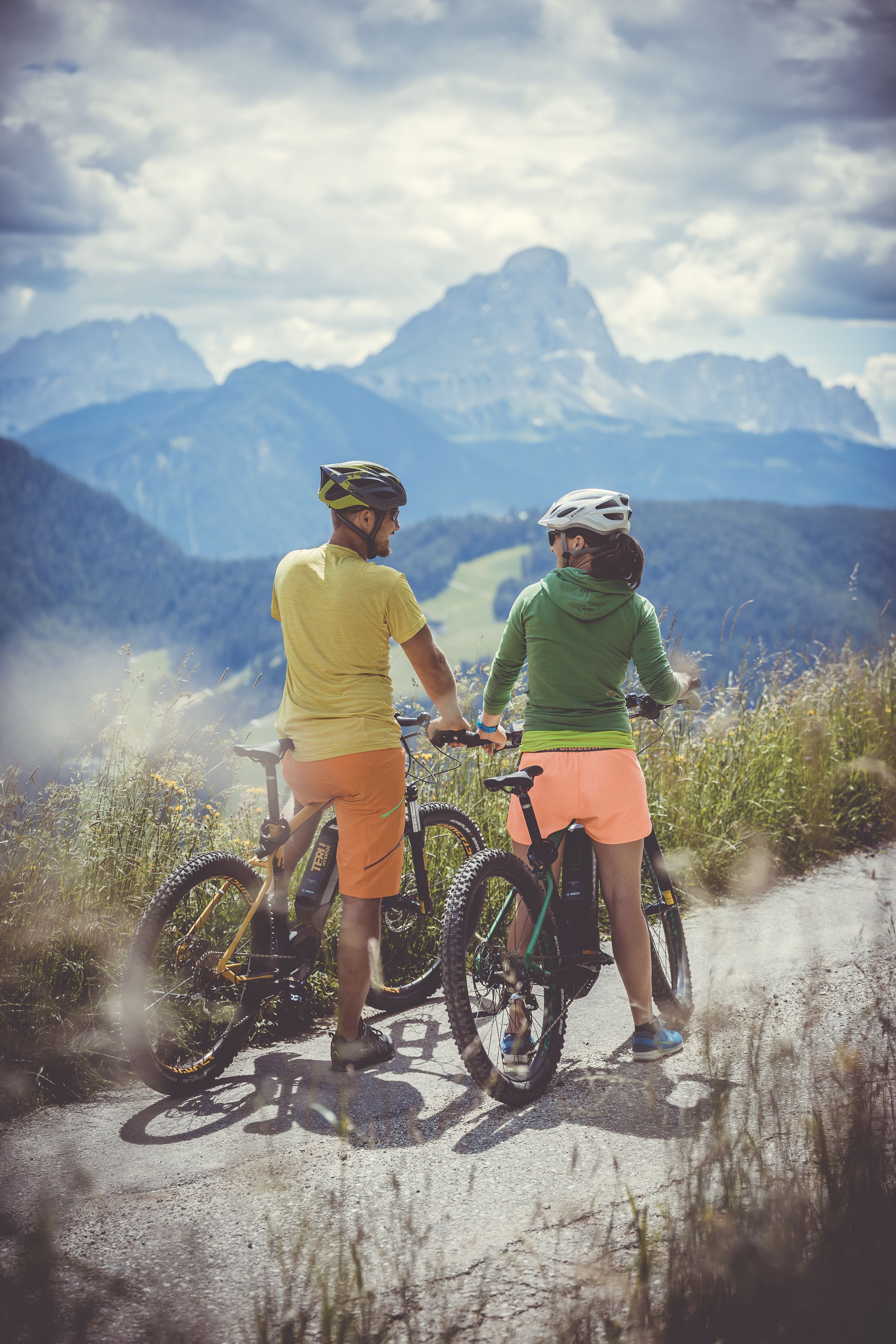 Two people on bicycles on a mountain path with a view of mountains in the background.