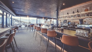 A modern bar with a high ceiling and exposed beams, featuring a bar counter with stools and shelves stocked with bottles and glasses.