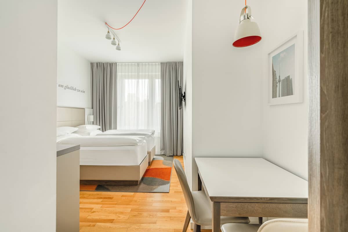 Une chambre d’hôtel avec un lit, un bureau et une chaise. Il y a une fenêtre avec des rideaux, une lampe fixée au mur et un tableau encadré accroché au mur.