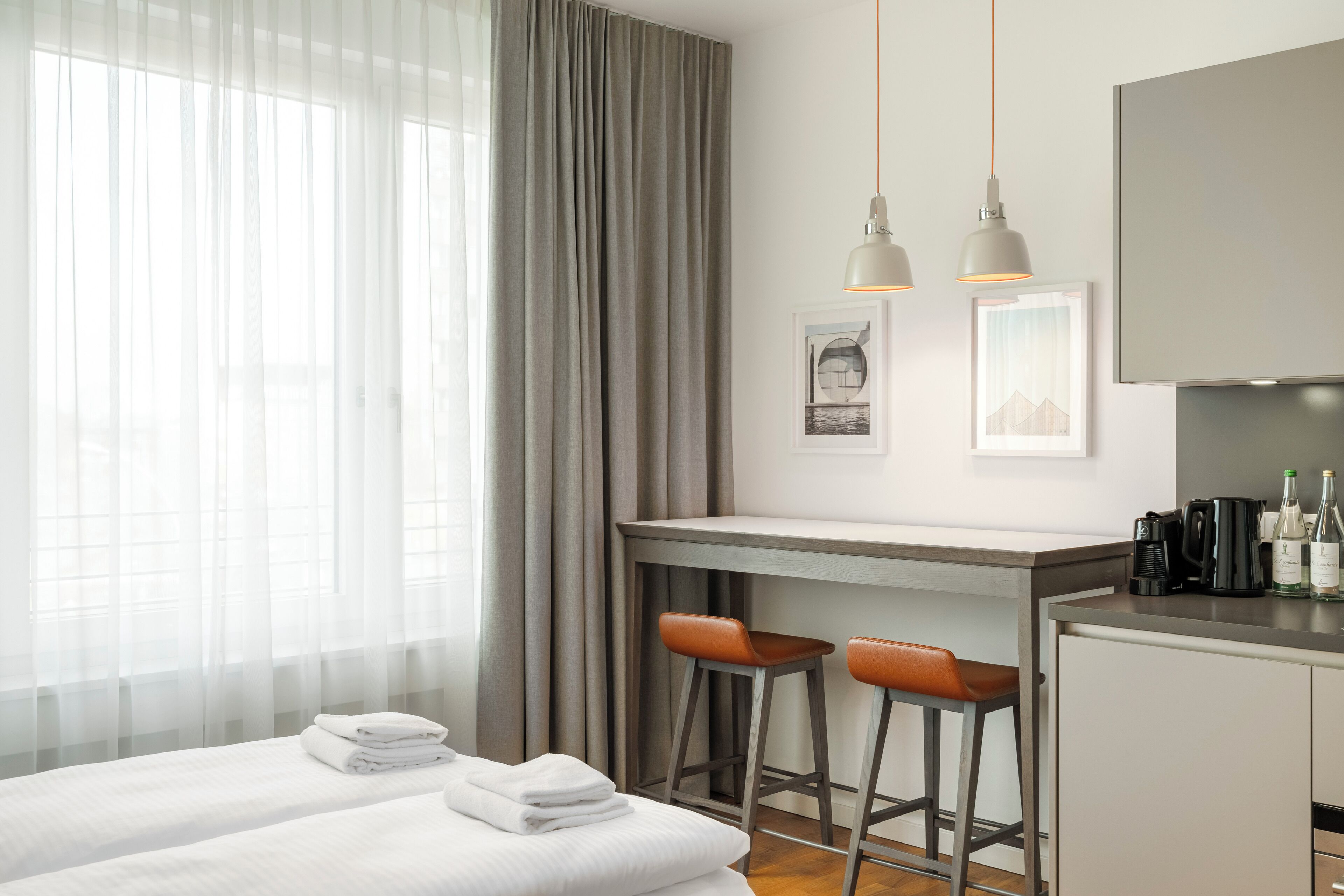 A hotel room with a bed, a desk, two bar stools, a coffee machine, and a minibar.