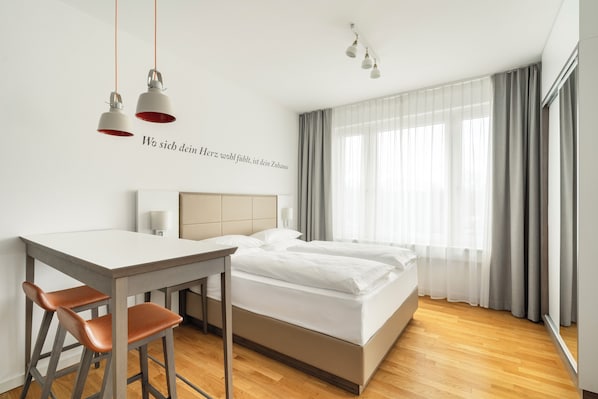 Une chambre d’hôtel moderne équipée d’un lit, d’un bureau, de deux tabourets de bar et d’une grande fenêtre avec des rideaux.