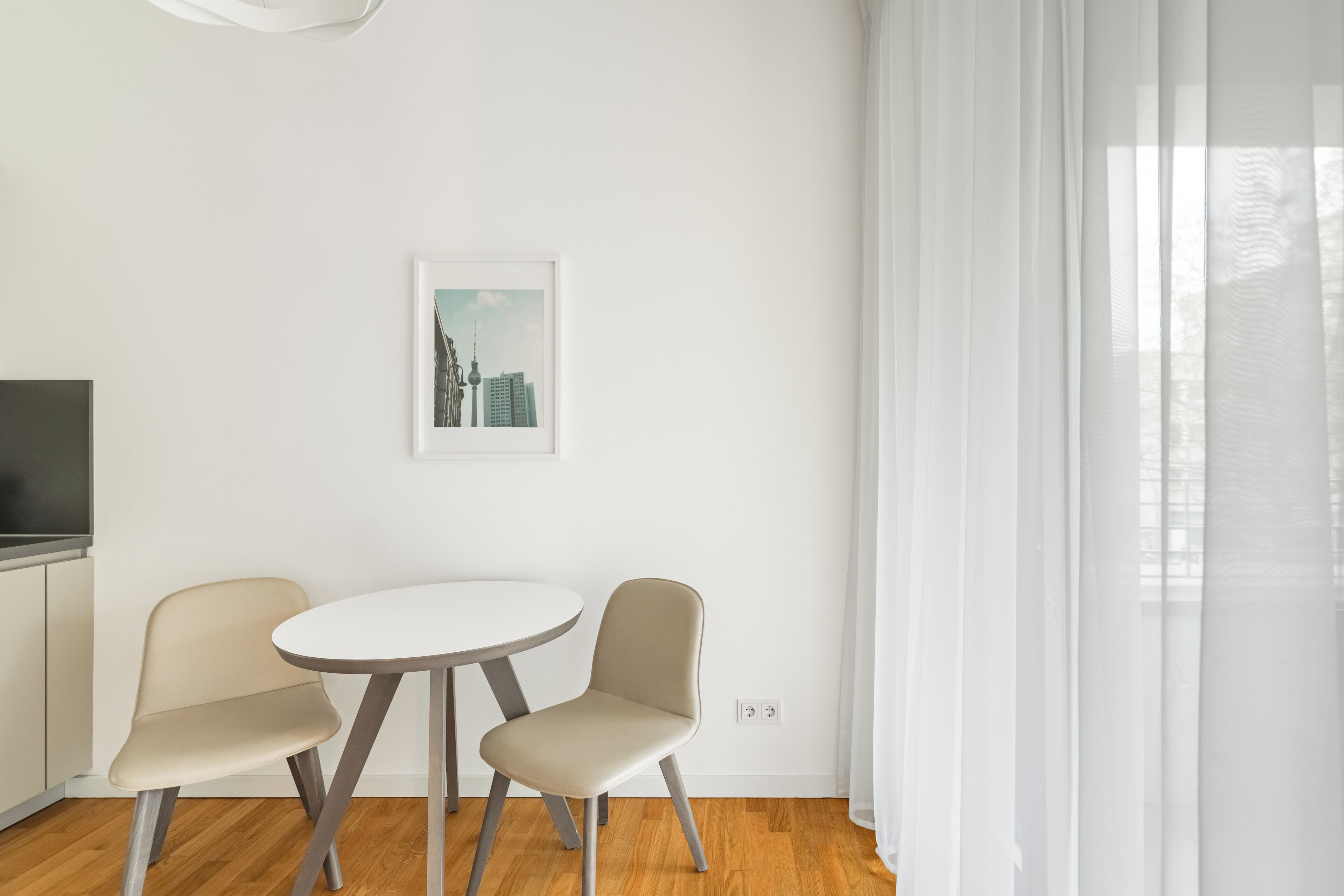 Una habitación moderna y minimalista con una mesa redonda y dos sillas, un cuadro con una vista urbana enmarcada en la pared, y un ventanal grande con cortinas translúcidas.