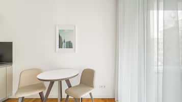 Una habitación moderna y minimalista con una mesa redonda y dos sillas, un cuadro con una vista urbana enmarcada en la pared, y un ventanal grande con cortinas translúcidas.