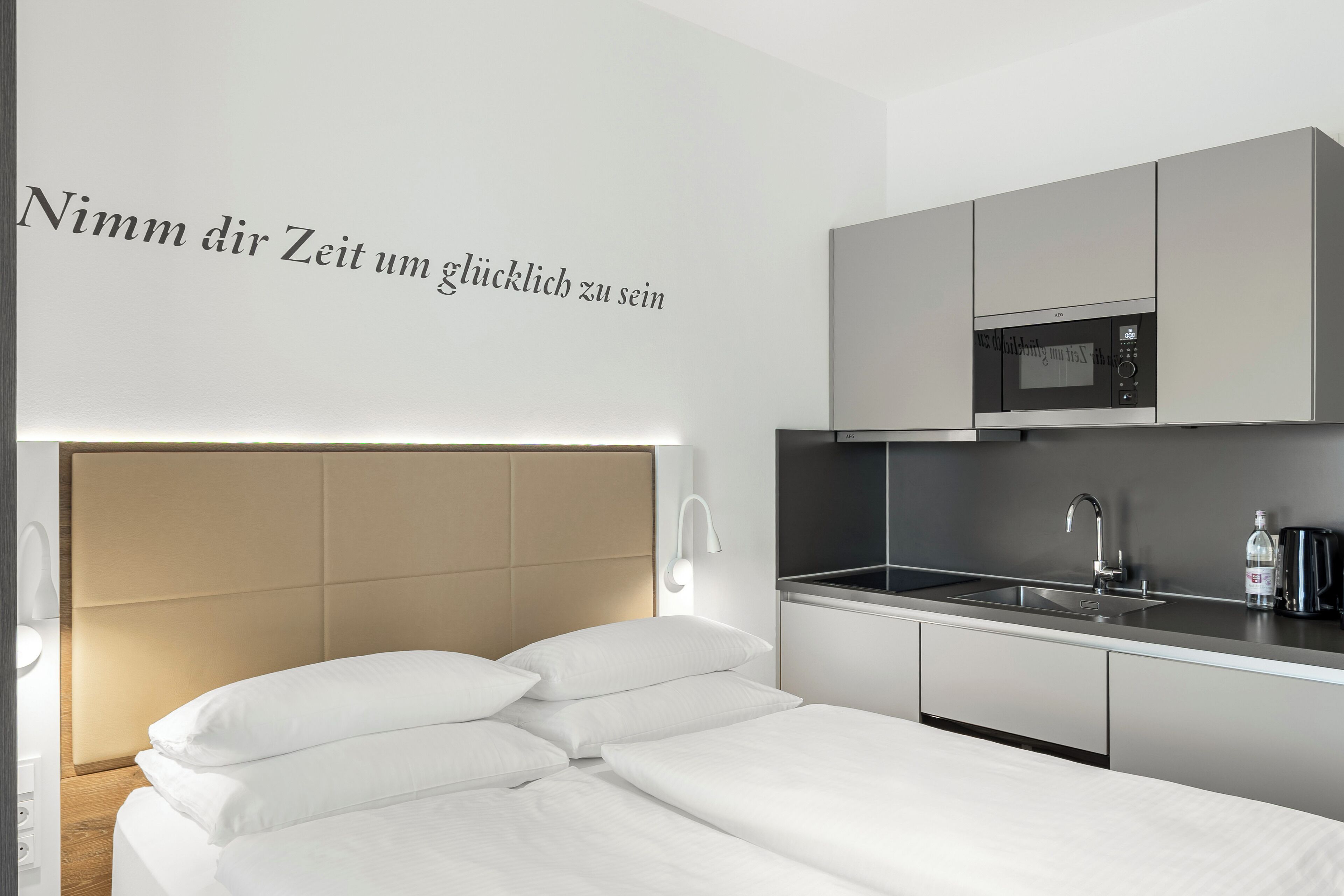 A modern bedroom with a bed, a bedside table, a wall-mounted light, and a kitchen area with a microwave and sink.