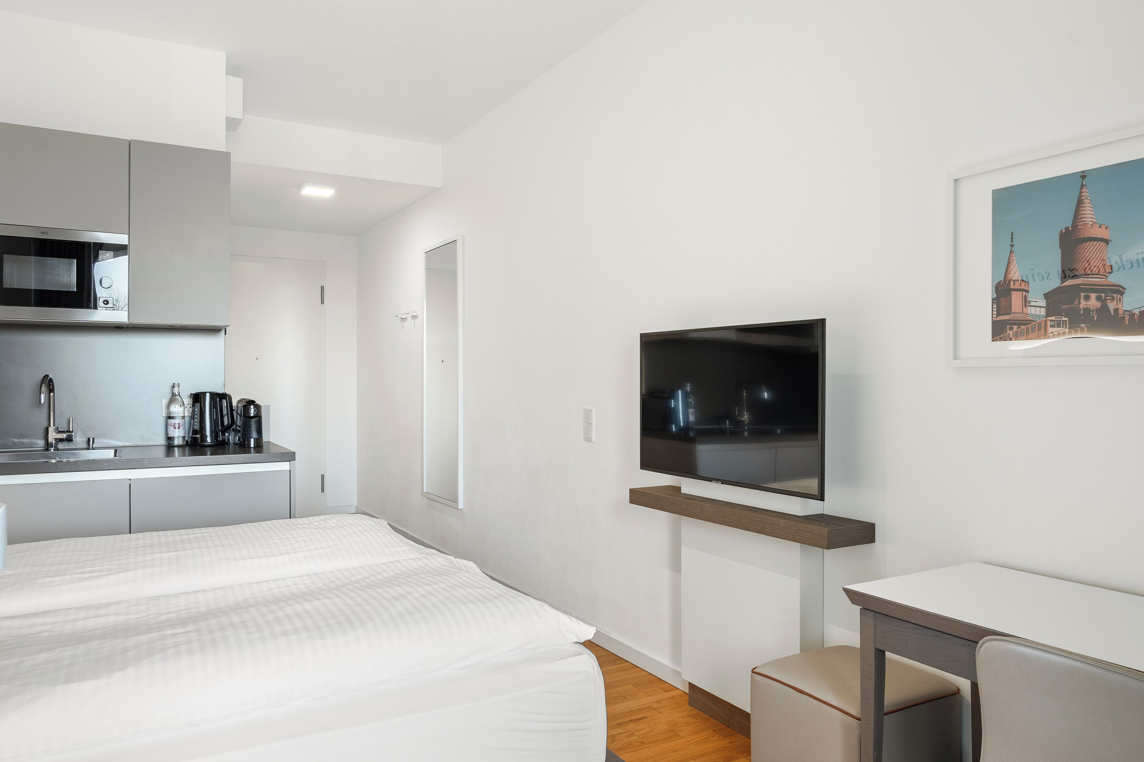 A modern hotel room with a bed, a small desk, a television, and a kitchenette.