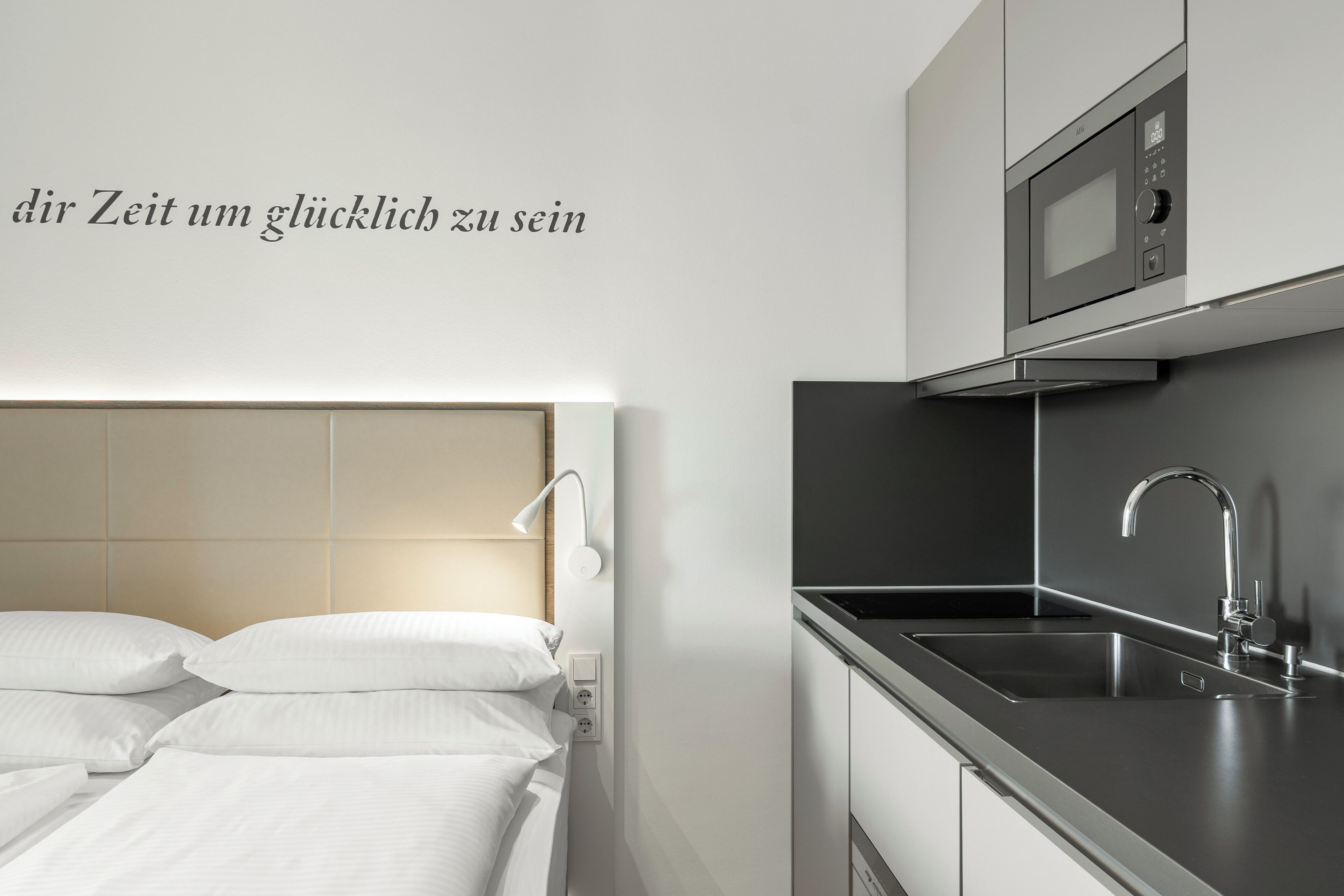 Una habitación de hotel moderna con cama, lámpara de noche y una kitchenette equipada con microondas y fregadero.