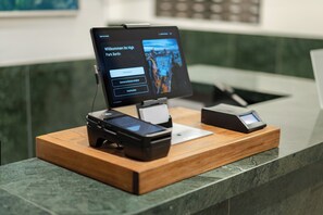 Un kiosque numérique doté d’un écran affichant un message d’accueil en allemand, d’un terminal de paiement et d’un lecteur de cartes, le tout installé sur un comptoir en bois.