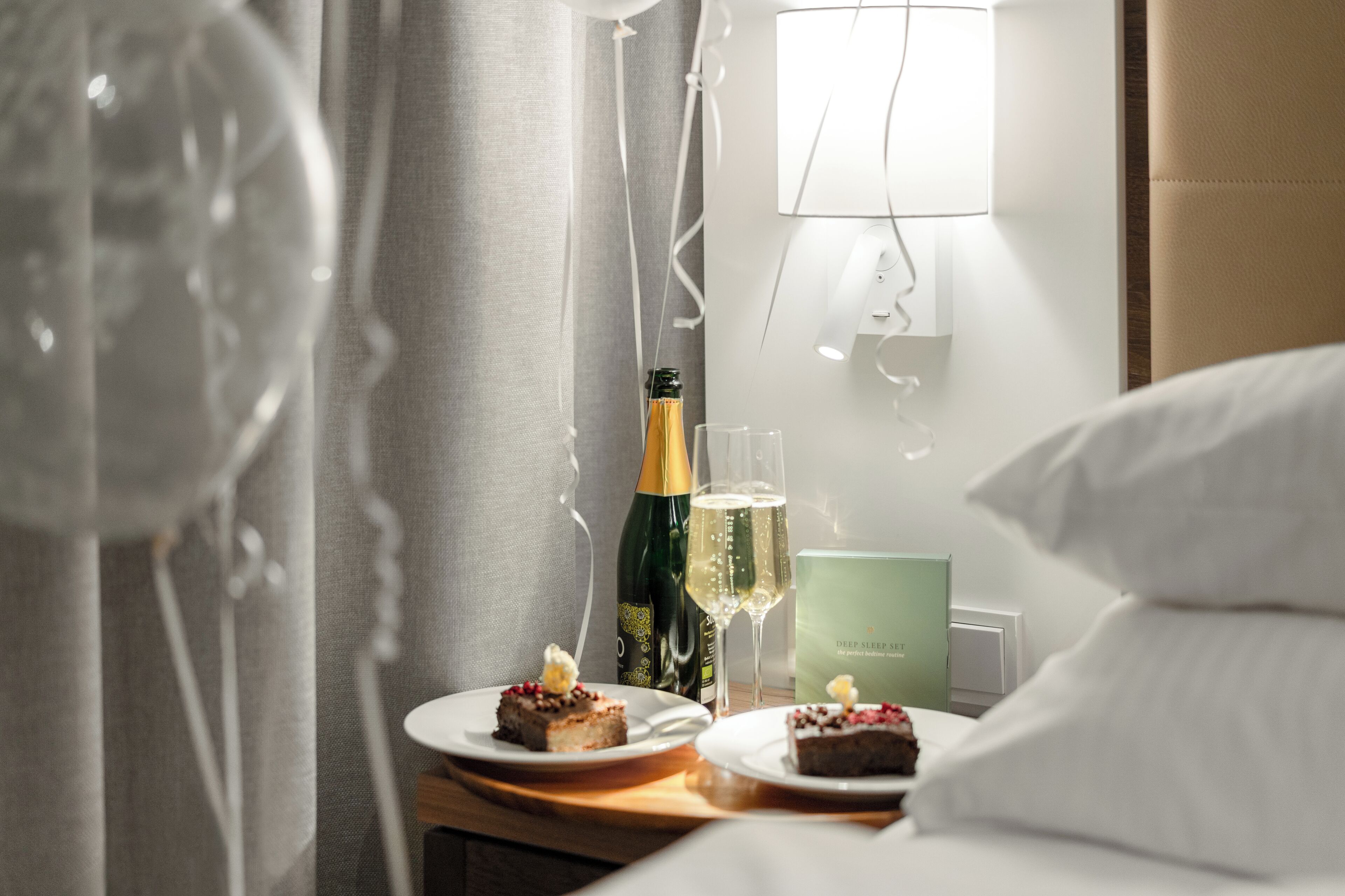 A hotel room with a bed, a nightstand with a bottle of champagne, two glasses of champagne, and two plates of dessert.