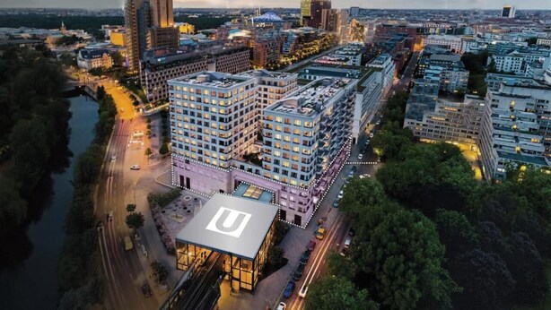 Un grand hôtel, composé de nombreux étages, avec un logo en forme de «U» bien visible sur le toit, entouré d’autres immeubles et bordé par une rivière.