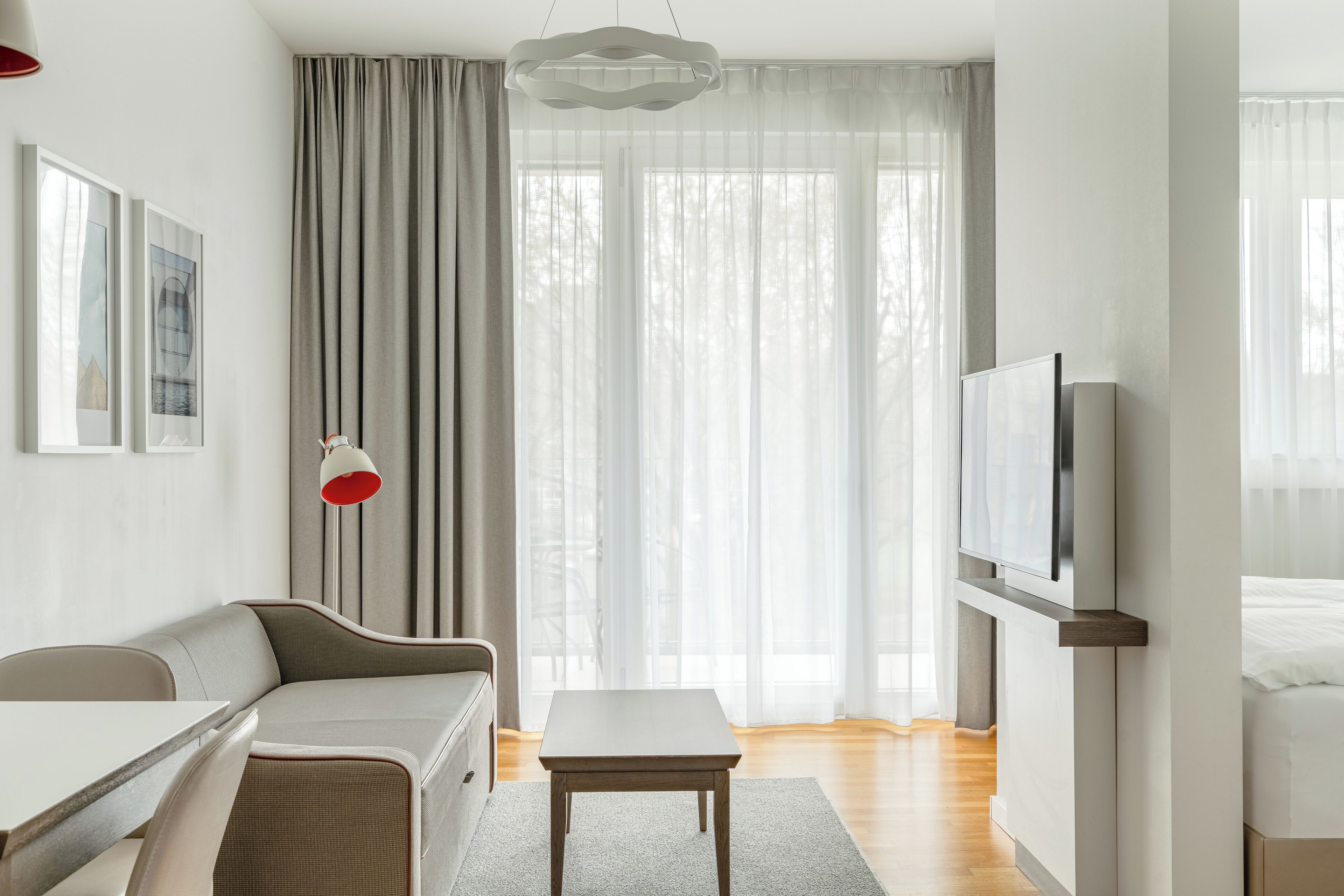 A modern hotel room with a sofa, a dining table, a TV, and large windows with sheer curtains.
