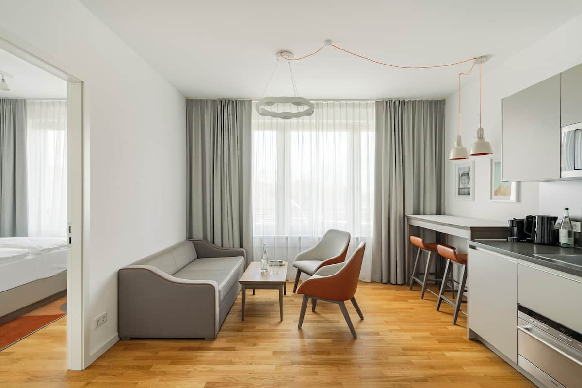 Une chambre d’hôtel moderne comprenant un canapé, deux fauteuils, une petite table, un coin cuisine avec un bar et une grande fenêtre avec des rideaux.