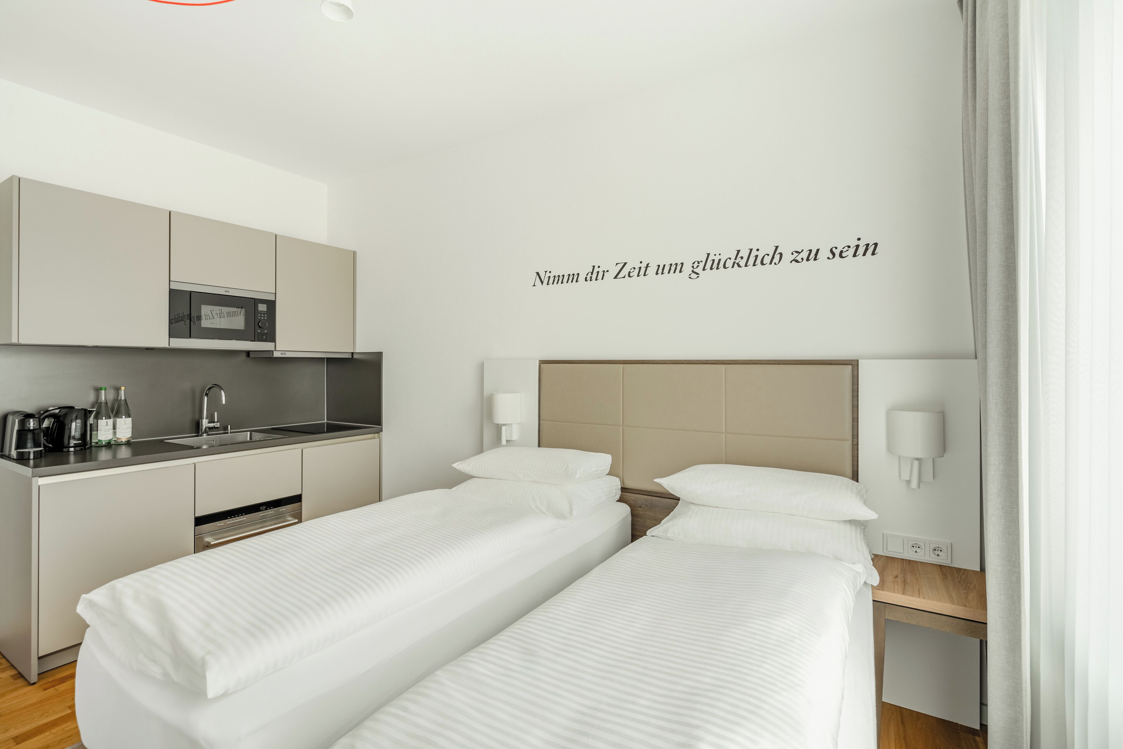 Een moderne hotelkamer met kitchenette, twee bedden en een wandlamp.
