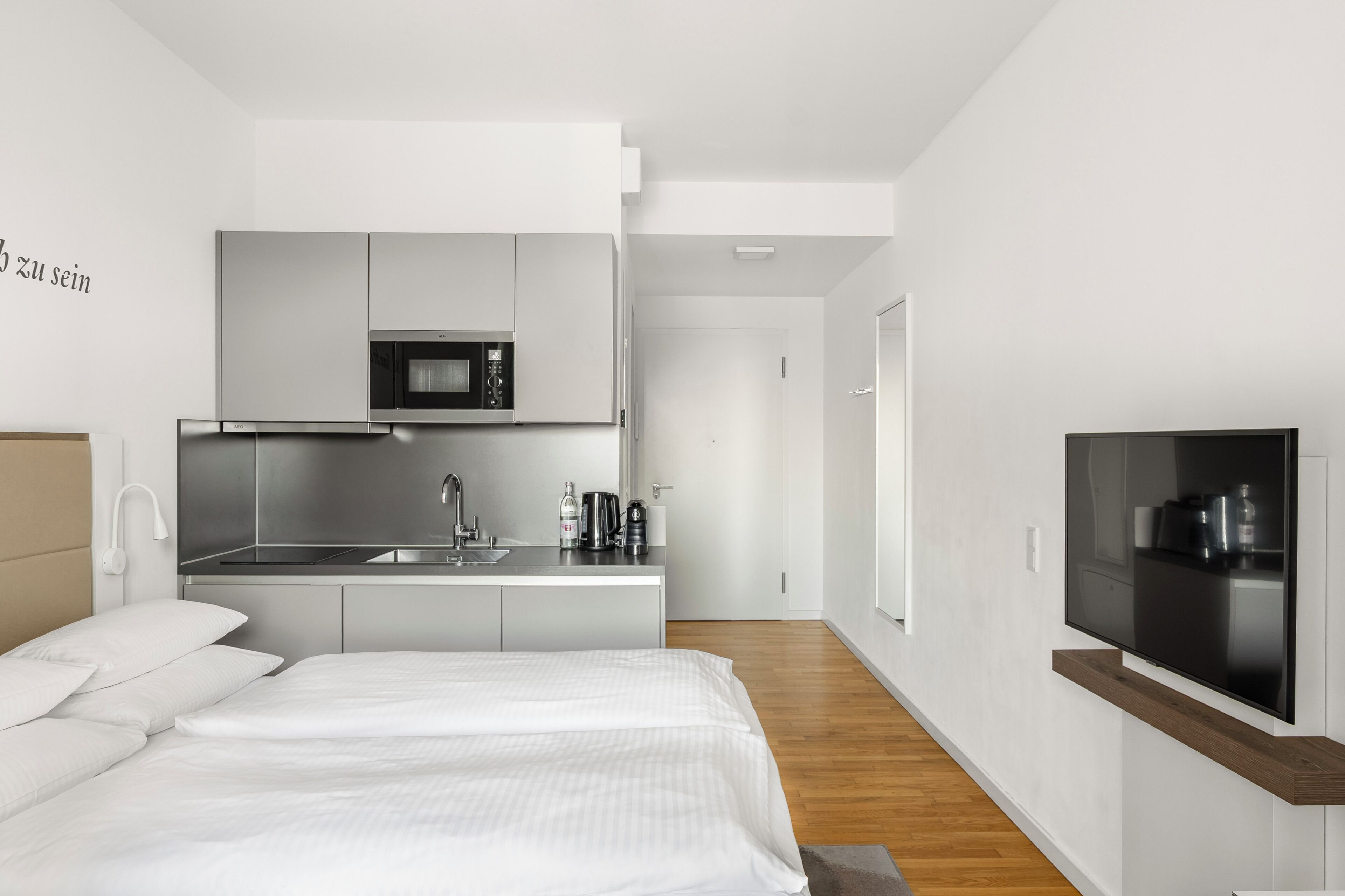 A modern hotel room with a bed, a kitchenette, and a TV.