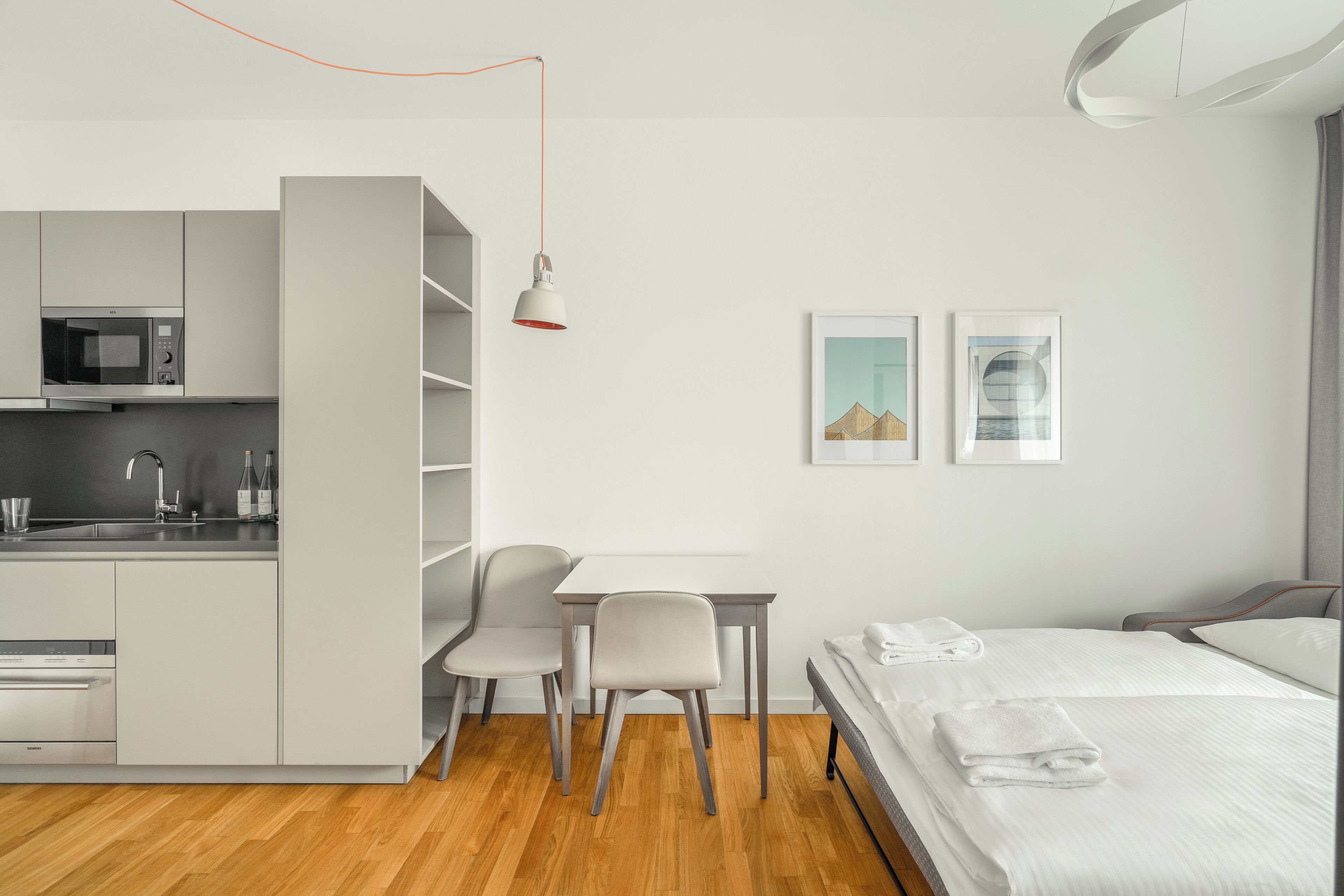 Habitación de hotel compacta con kitchenette, una cama con toallas ordenadas, un escritorio con silla y dos cuadros en la pared.