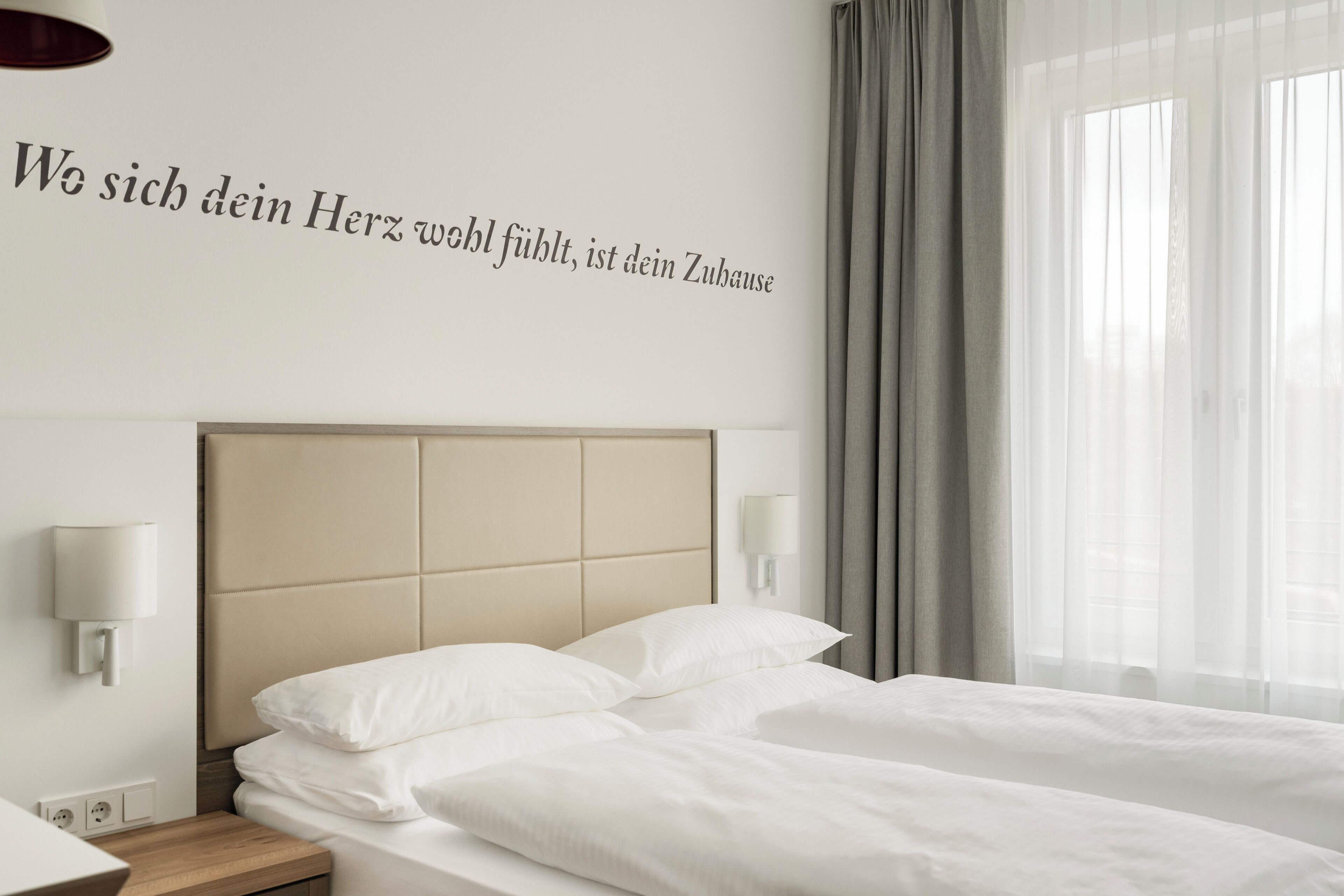 Una habitación de hotel con una cama, cabecera, lámpara de pared y ventana con cortinas.