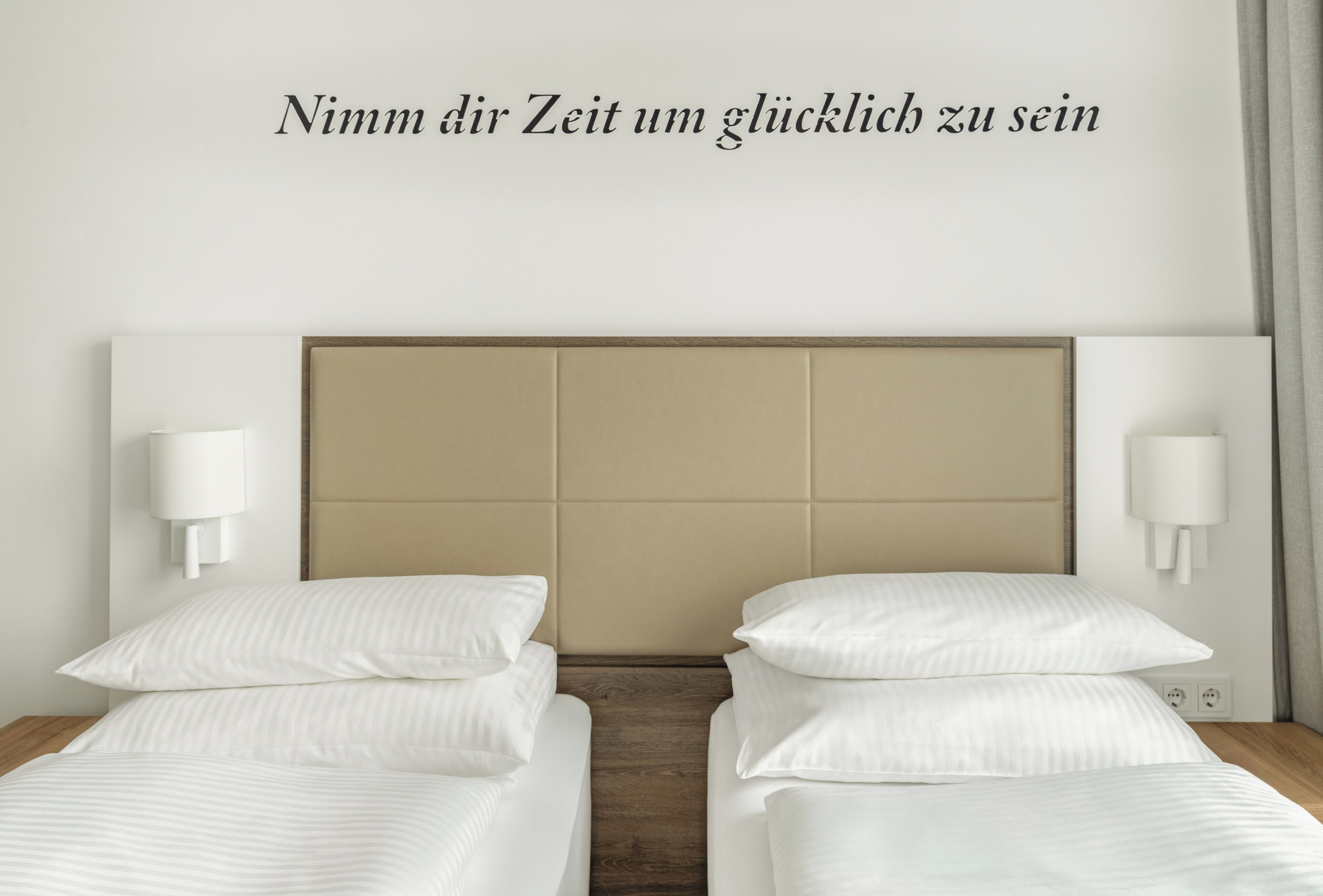 Habitación de hotel con dos camas, un cabecero color beige, ropa de cama blanca y lámparas fijadas a la pared.