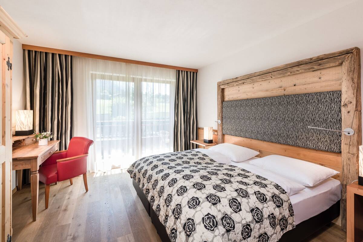 Une chambre d’hôtel avec une tête de lit en bois, un lit recouvert d’une courtepointe à motifs, un bureau avec une lampe, une chaise rouge et une grande fenêtre avec des rideaux.