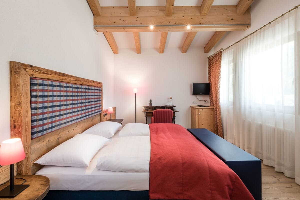 Une chambre avec une tête de lit en bois, un lit avec une literie blanche et rouge, un bureau avec une chaise, une télévision et une grande fenêtre avec des rideaux.