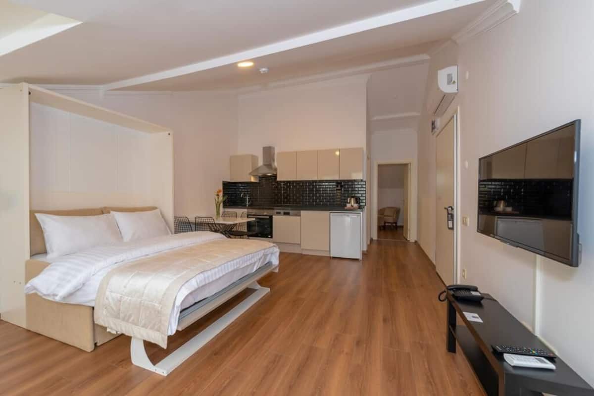 A modern hotel room with a bed, a desk, a TV, and a kitchenette.