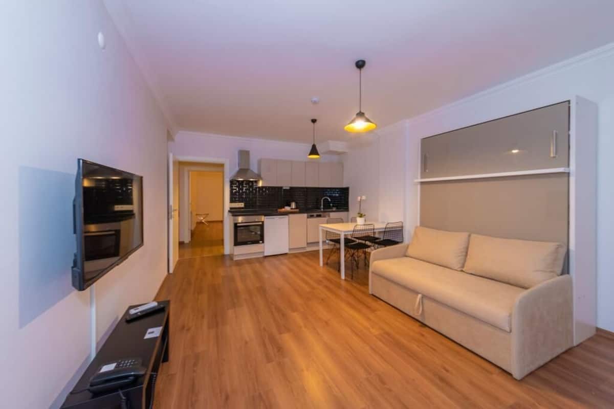 A modern living room with a sofa, a TV mounted on the wall, a dining table with chairs, and a kitchen area with cabinets and a stove.