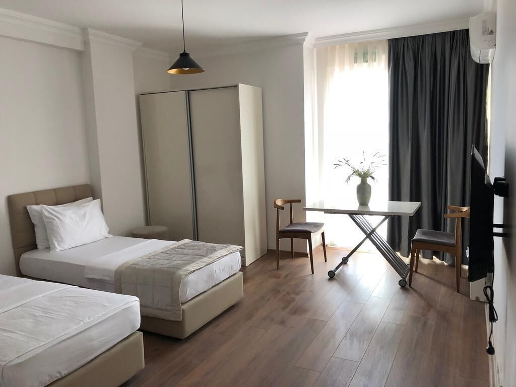 A hotel room with two beds, a desk, a chair, a potted plant, and a window with curtains.