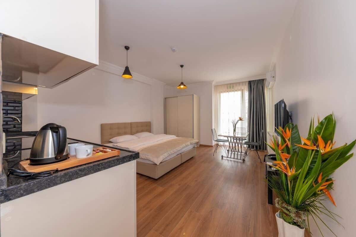A modern hotel room with a bed, kitchenette, dining area, and a large window.