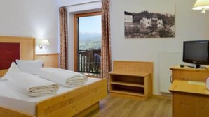 A hotel room with a wooden bed, a television, a desk, and a window with a view of a mountain landscape. - Hotel Haus an der Luck (Barbiano)