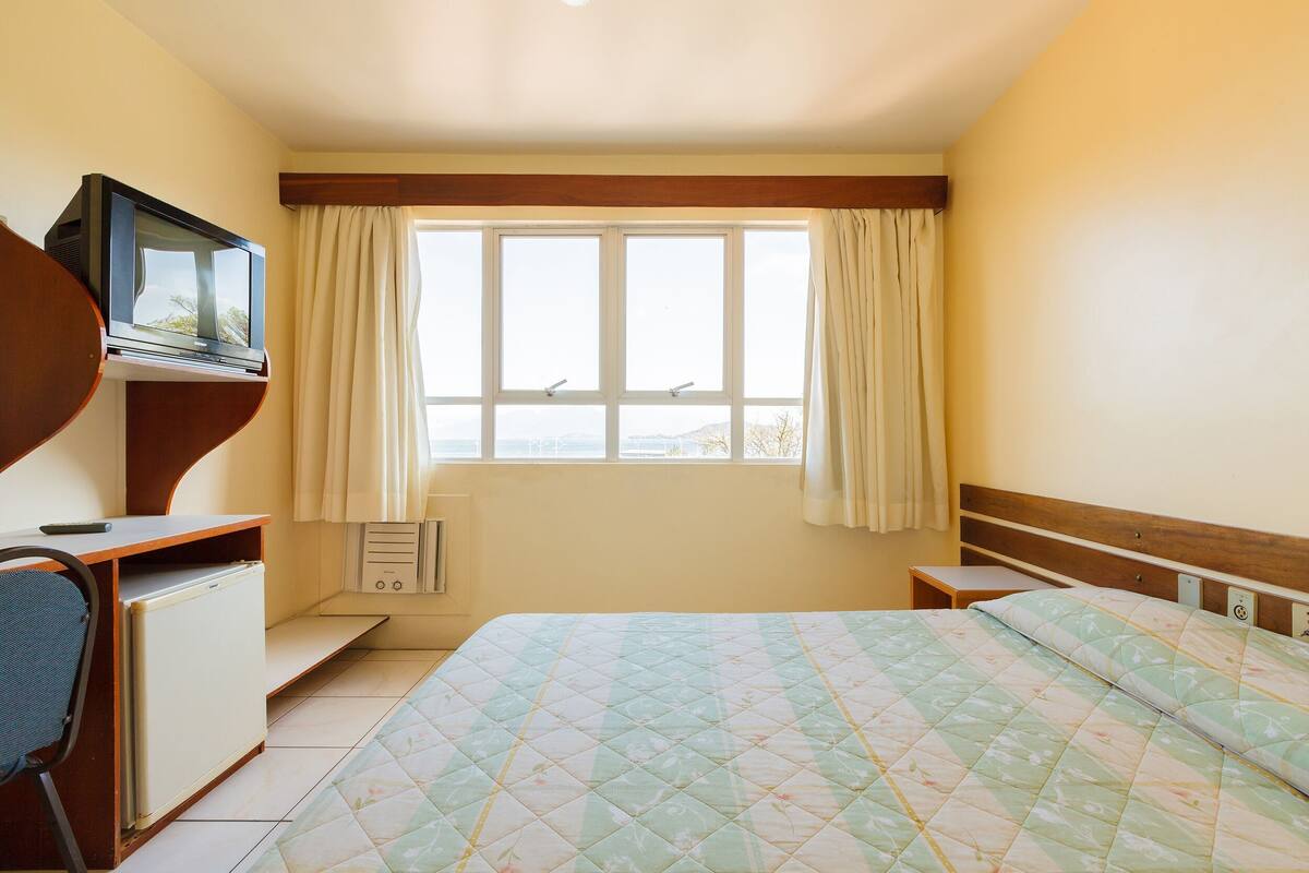 A bedroom with a bed, a TV on a stand, a desk with a chair, and a window with curtains.