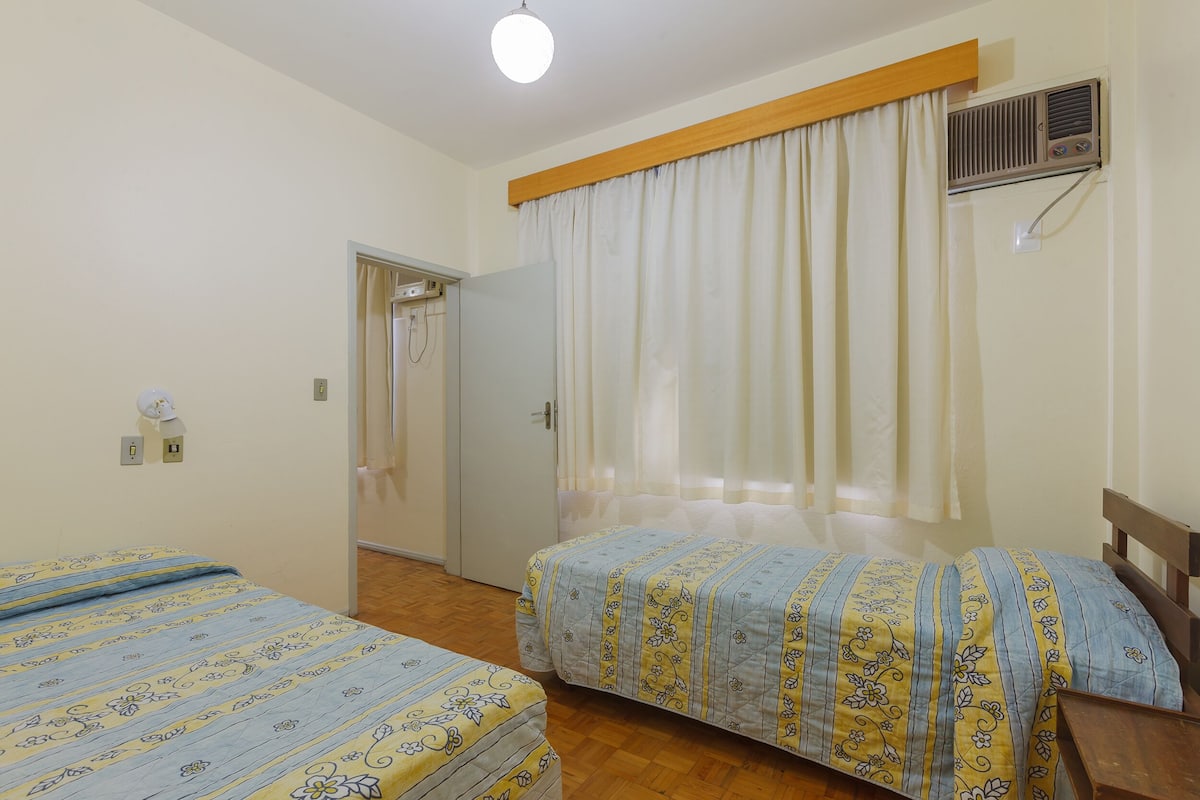 A bedroom with two beds, a wooden headboard, a ceiling fan, a window with curtains, and a door leading to another room.