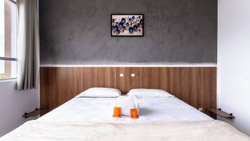Quarto de hotel moderno com cabeceira de madeira, uma cama grande com roupa de cama branca, uma obra de arte emoldurada acima da cama e uma janela com cortinas.