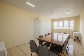 A conference room with a large wooden table, eight chairs, a door with a 'SAÍDA' sign, a window with a city view, and a small table with a white tablecloth. - Tri Hotel Florianópolis (Florianópolis)